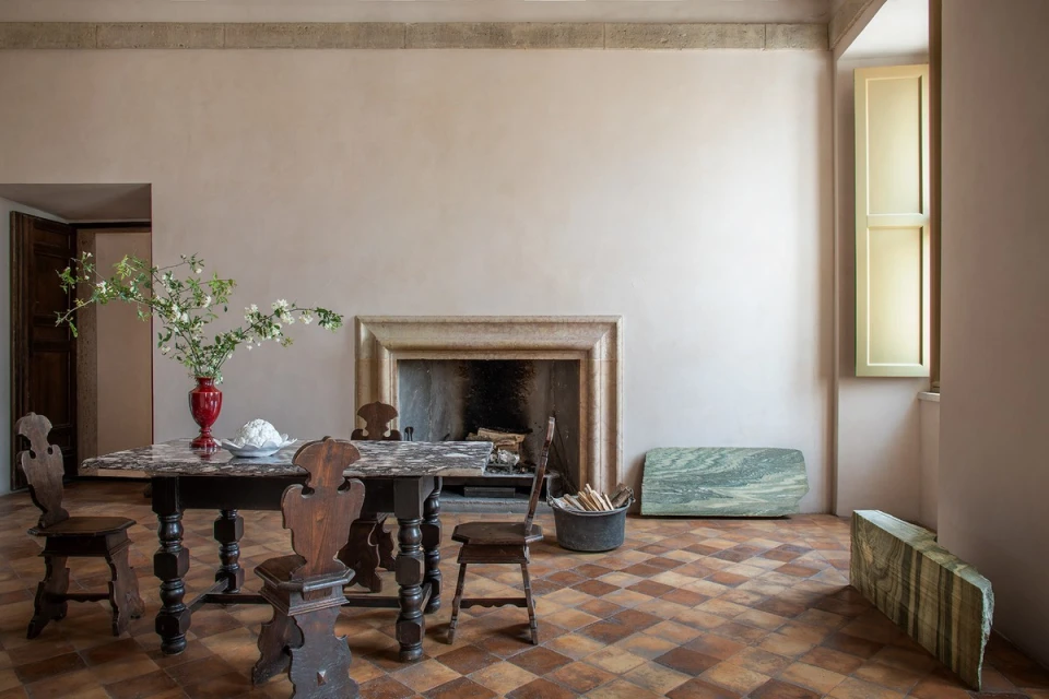 Indoor shot showcases a dark wooden table with chairs, a red vase with flowers, a fireplace, and checkered tile flooring.