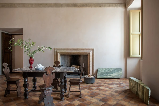 Indoor shot showcases a dark wooden table with chairs, a red vase with flowers, a fireplace, and checkered tile flooring.