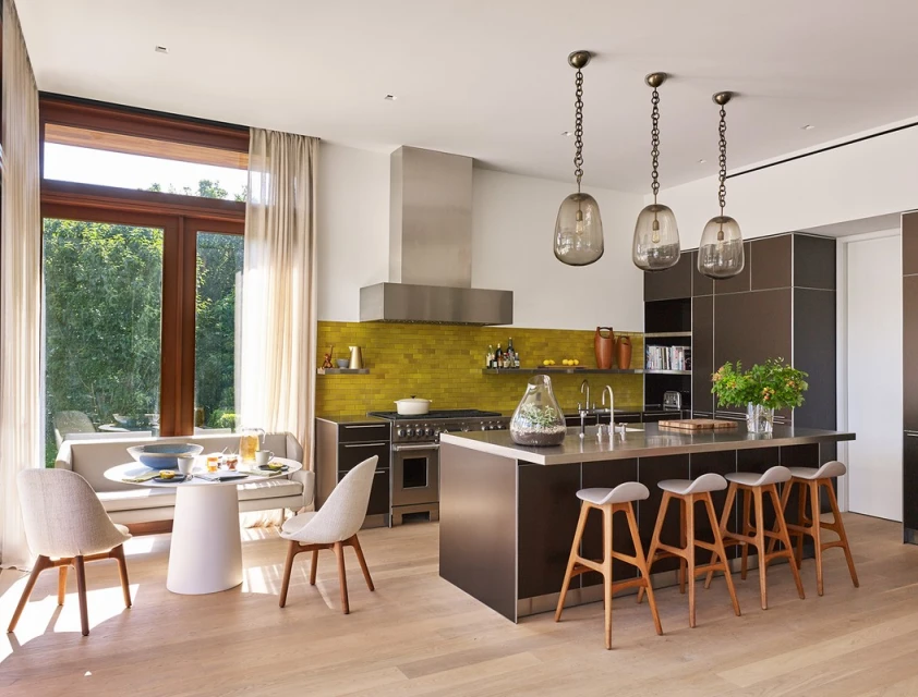 Bright and modern kitchen interior featuring a central island with bar stools, a dining area with table and chairs, and large windows.