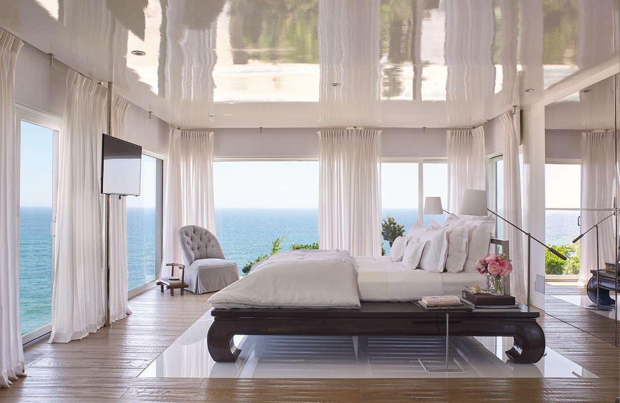 Bedroom with ocean view features a large bed, tufted chair, floor-to-ceiling windows with white curtains, and reflective ceiling.