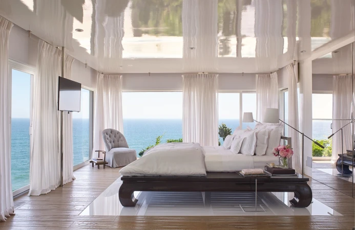 Bedroom with ocean view features a large bed, tufted chair, floor-to-ceiling windows with white curtains, and reflective ceiling.