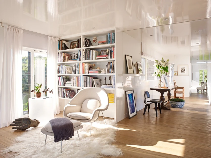 Bright and airy living room with a white chair, bookshelf, and wooden floor. Natural light fills the interior space.