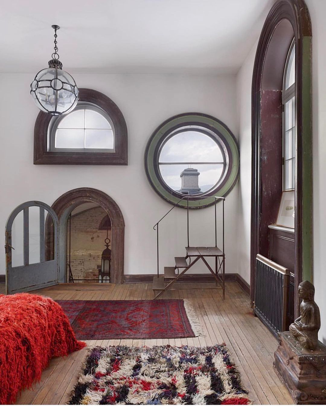 Photo of a bedroom featuring multiple arched windows, a bed with a shaggy red cover, oriental rugs, and a Buddha statue.