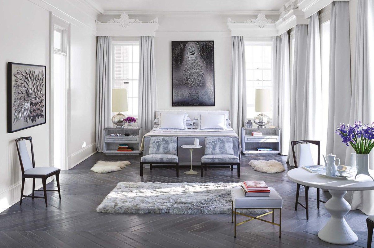 Well-lit bedroom featuring a bed with white pillows, matching benches, bedside tables with lamps, and a round table with chairs.