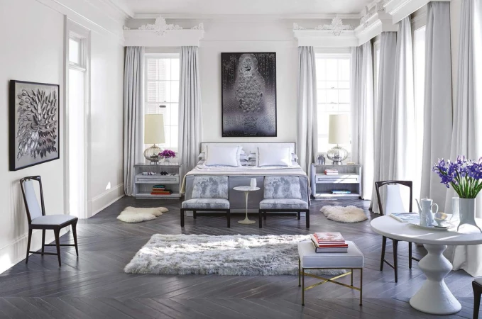Well-lit bedroom featuring a bed with white pillows, matching benches, bedside tables with lamps, and a round table with chairs.
