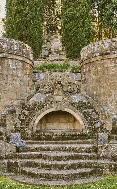 Stone fountain with a large face carved in it. There are stone steps leading up to the fountain, surrounded by trees in the background.