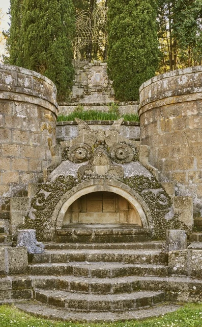 Stone fountain with a large face carved in it. There are stone steps leading up to the fountain, surrounded by trees in the background.