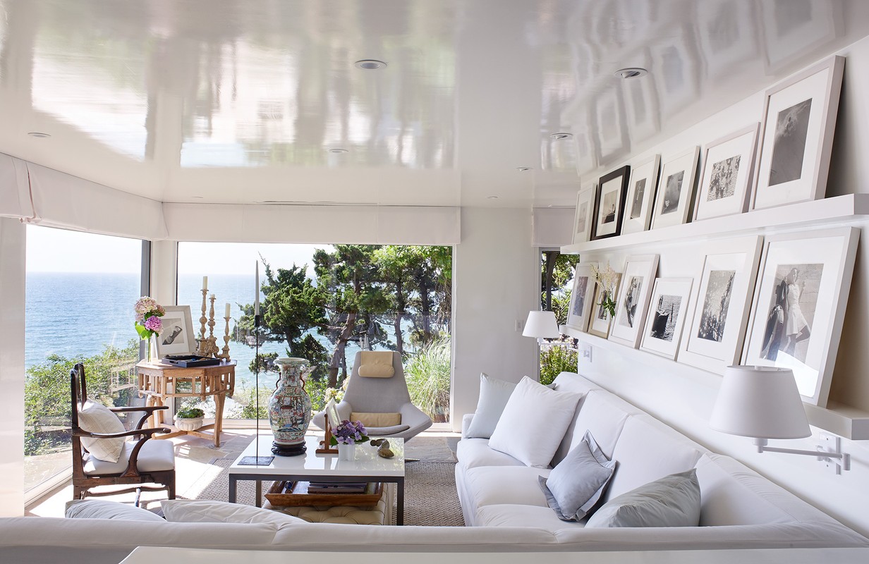 Bright living room interior with white furniture, seascape views, and gallery wall of framed black-and-white photographs creating a cozy atmosphere.