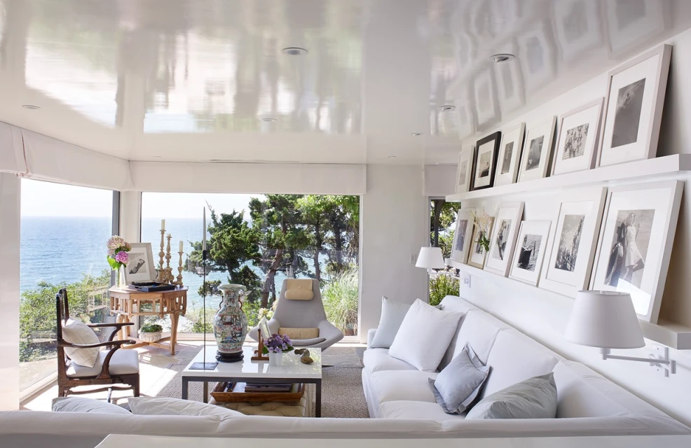 Bright living room interior with white furniture, seascape views, and gallery wall of framed black-and-white photographs creating a cozy atmosphere.
