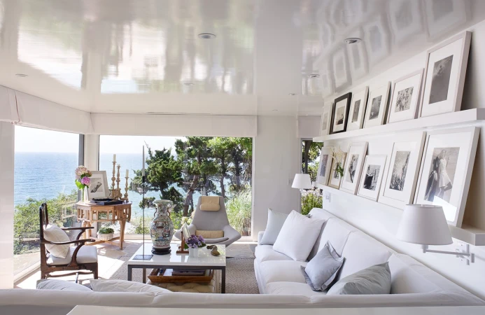 Bright living room interior with white furniture, seascape views, and gallery wall of framed black-and-white photographs creating a cozy atmosphere.