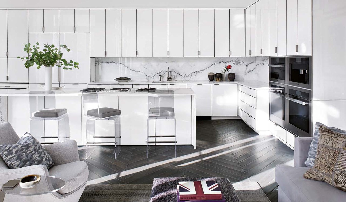 Modern kitchen interior with white cabinets, island with stools, herringbone floor, gray sofa and chairs and natural light.
