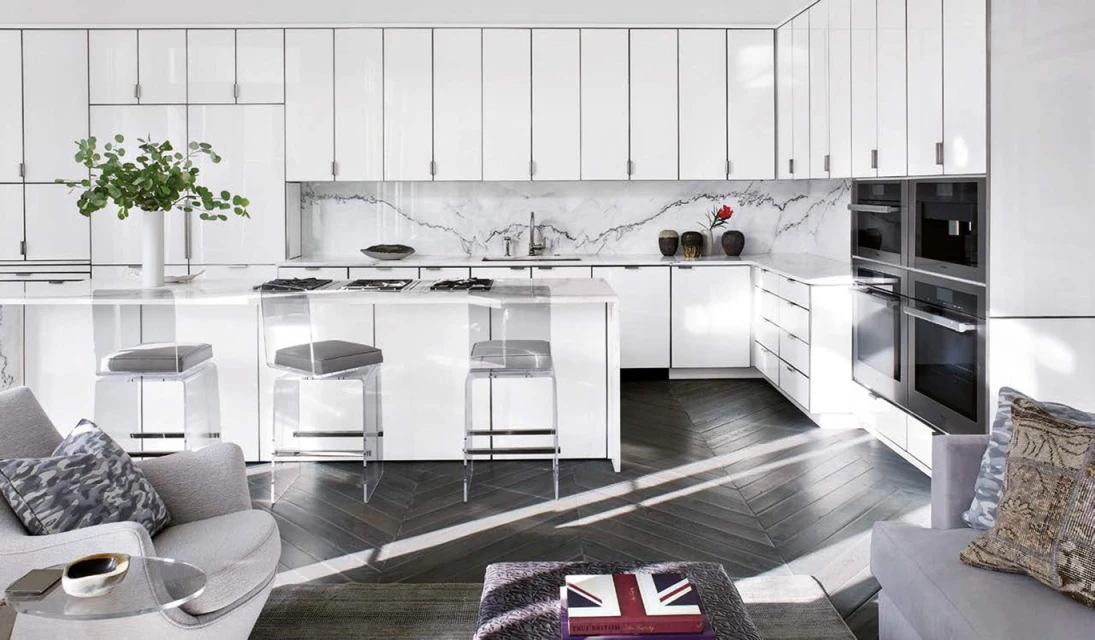 Modern kitchen interior with white cabinets, island with stools, herringbone floor, gray sofa and chairs and natural light.