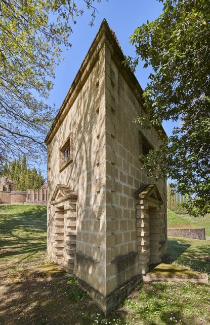 Stone building exterior with symmetrical design and arched doorways surrounded by a green grass lawn and trees under a clear blue sky.