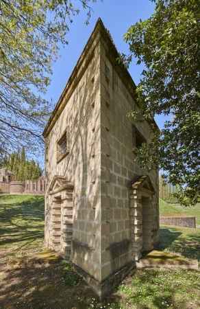 Stone building exterior with symmetrical design and arched doorways surrounded by a green grass lawn and trees under a clear blue sky.