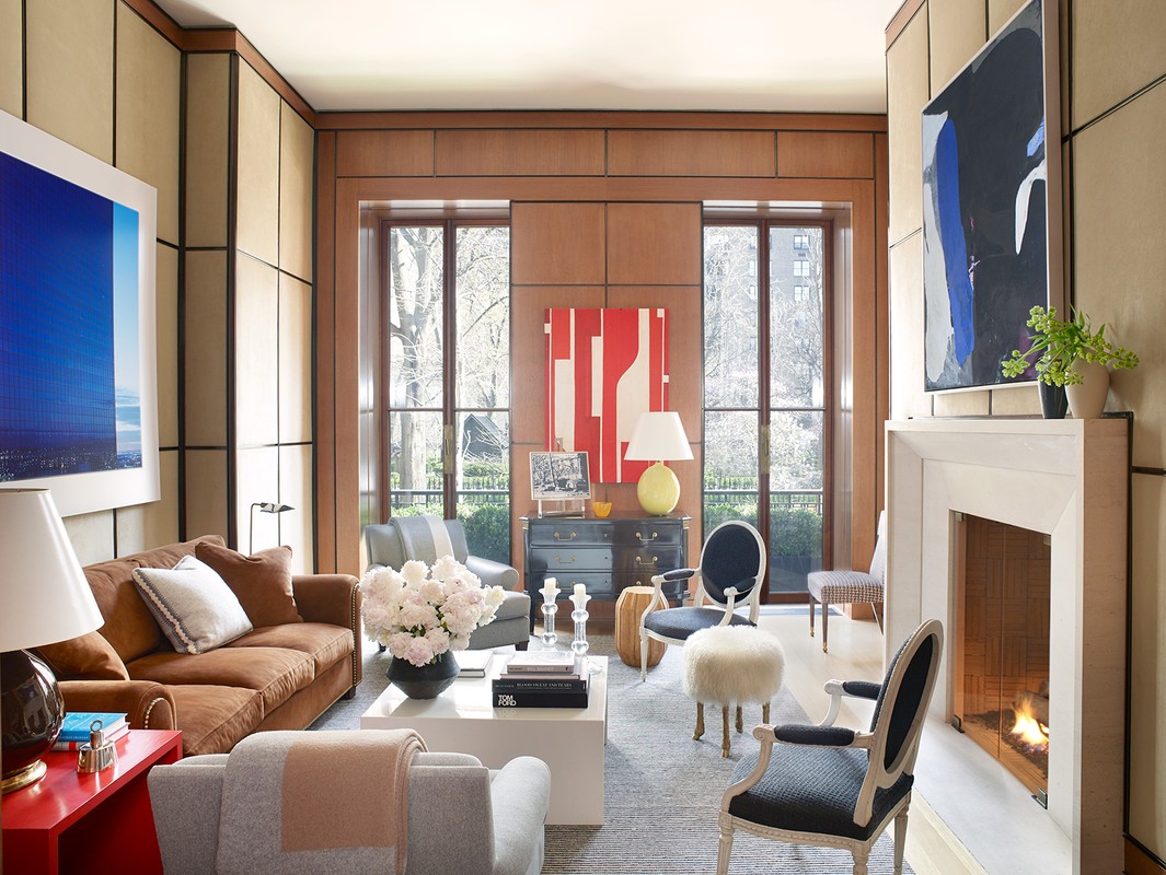 Living room interior with a brown sofa, white coffee table, chairs, fireplace, and artwork, bathed in natural light from large windows.