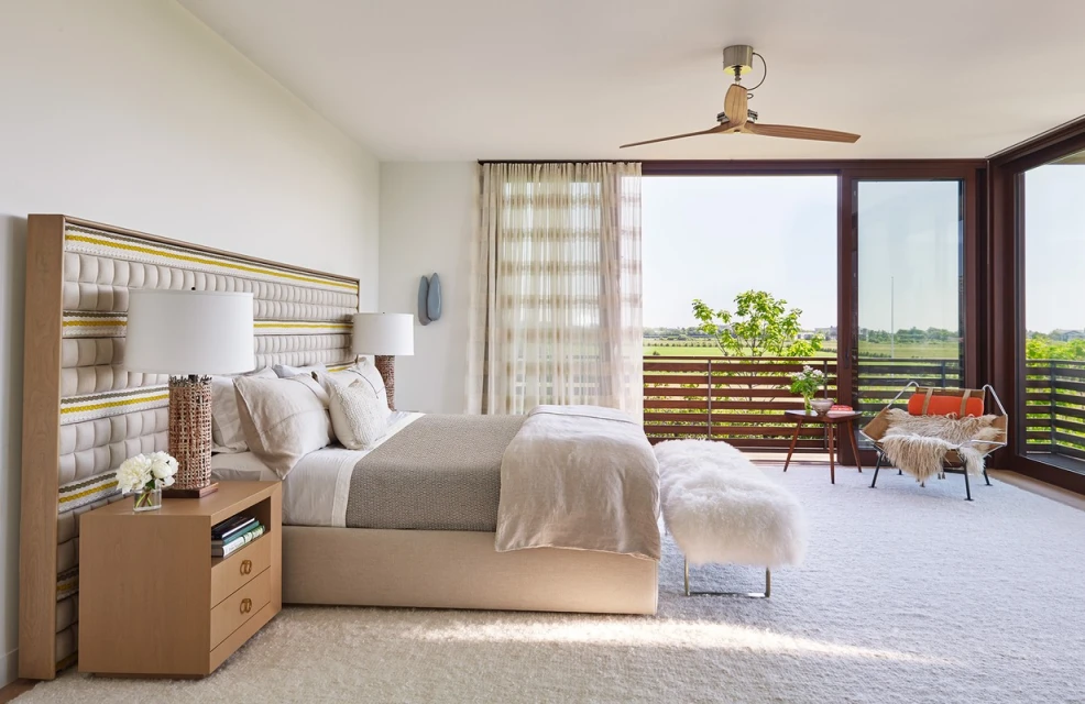 Bedroom interior with large upholstered headboard, nightstand with lamps, plush carpet, and a balcony overlooking a landscape view.