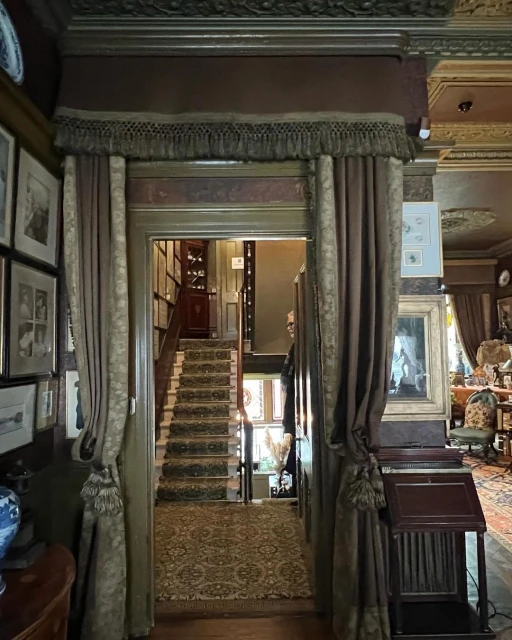 Elegant home interior featuring a hallway with a staircase, ornate decor, curtains, and vintage-style furnishings, creating a warm, inviting atmosphere.