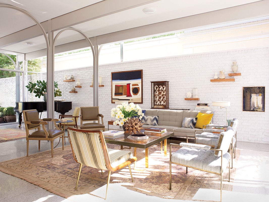 Living room interior featuring neutral-toned seating, coffee table with books and flowers, brick walls, and architectural arch details.