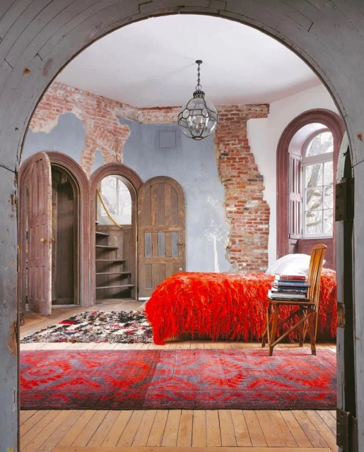 Eclectic bedroom interior featuring a red bed, arched doors, exposed brick wall, patterned rugs, and a vintage chandelier.