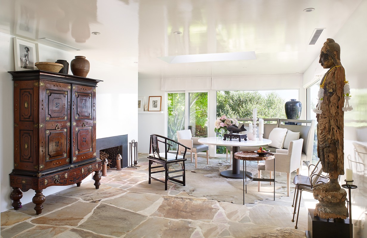 Eclectic dining room with a vintage cabinet, stone floors, sculpture, and a round table with flowers in an indoor setting.