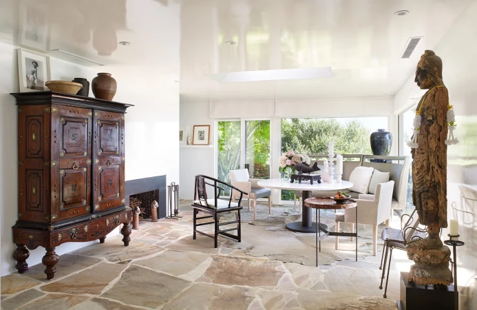 Eclectic dining room with a vintage cabinet, stone floors, sculpture, and a round table with flowers in an indoor setting.