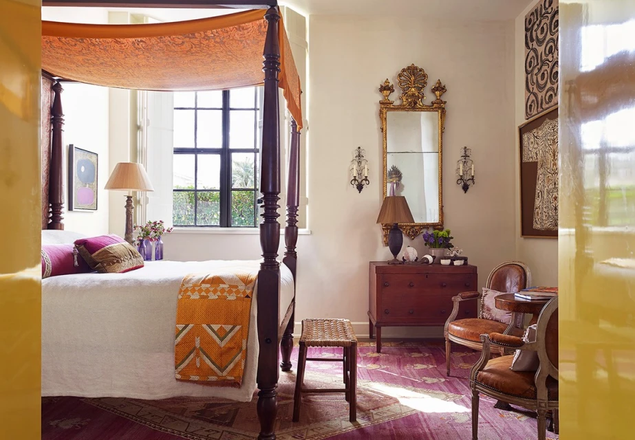 Bedroom interior with a four-poster bed, a wooden dresser, decorative mirror, and seating creating a warm and inviting space.