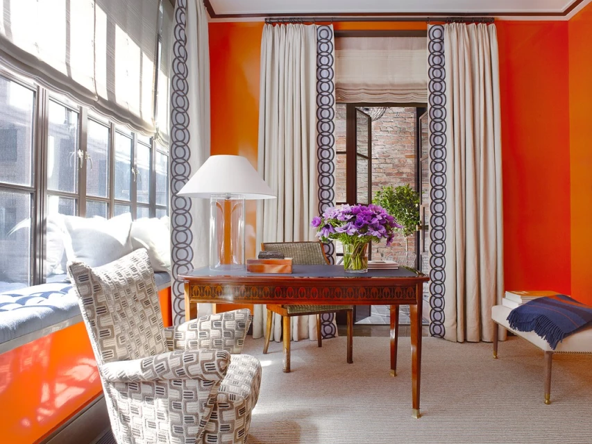 Bright interior space with orange walls, featuring a desk with flowers, patterned chair, window seat, and neutral-toned carpet flooring.