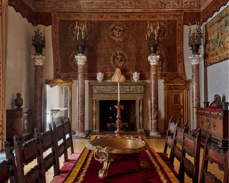 Ornate dining room with long table set with gold bowl, flanked by carved wood chairs and marble columns. Decorative tapestries hang.