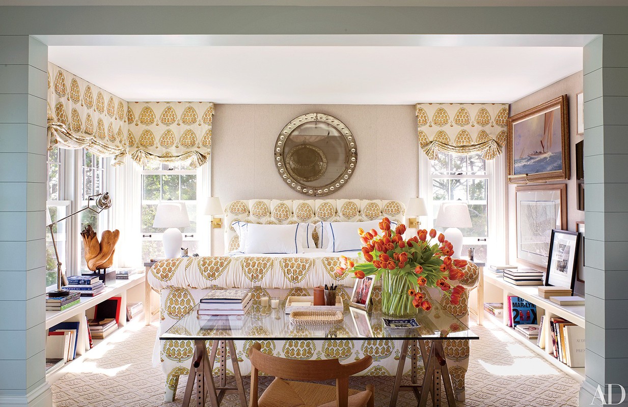 Bedroom interior with a glass-topped desk, floral-patterned headboard, window seating with patterned valances, and a round mirror.