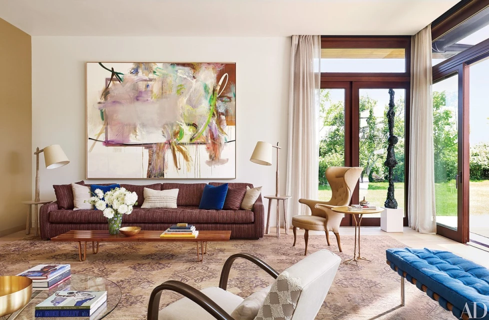 Living room interior featuring a brown sofa with throw pillows, artwork, chairs, coffee tables, and a patterned rug in natural light.
