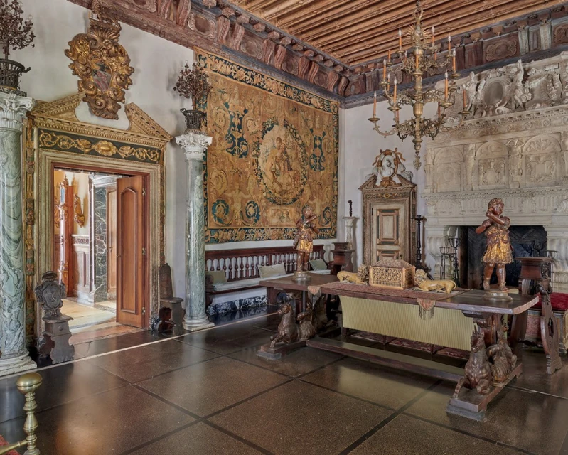 Ornate interior with a long wooden table topped with objects, flanked by statues, in a large room with a decorative tapestry.