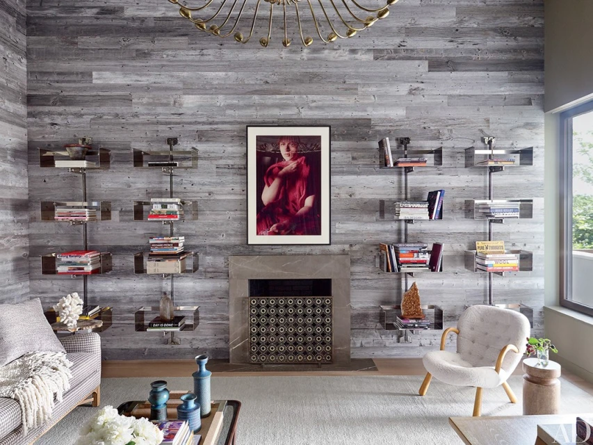 Living room interior with gray wood paneling, bookshelves, a fireplace, area rug, artwork, and a white upholstered chair.