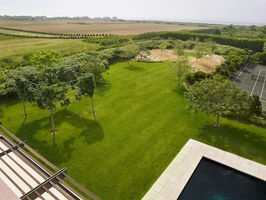 Overhead landscape showcases a manicured lawn with shade trees, a tennis court, a black swimming pool, and an open field under daylight.