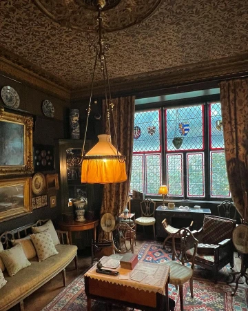Interior shot of a richly decorated historical room with dark walls, antique wooden furniture, patterned rugs, and stained glass windows.