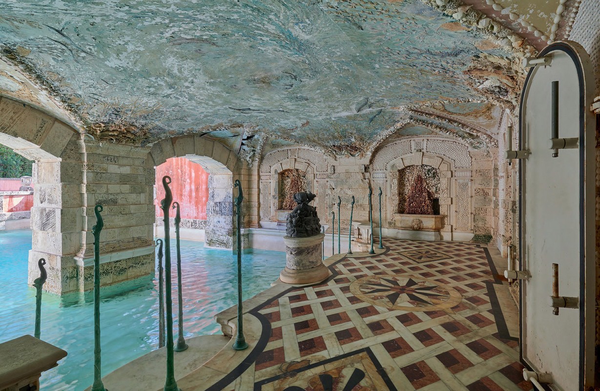 Ornate indoor grotto with turquoise pool, stone arches, shell-encrusted ceiling, and tiled floor. Natural light filters in from outside.