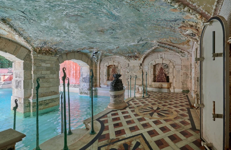Ornate indoor grotto with turquoise pool, stone arches, shell-encrusted ceiling, and tiled floor. Natural light filters in from outside.