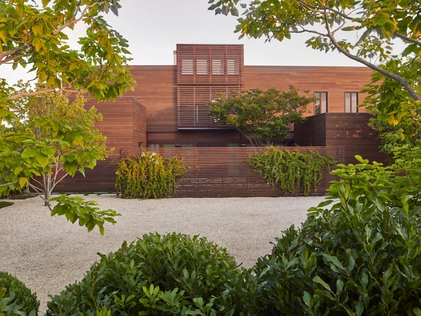 Exterior of a modern wood-clad house with gravel landscaping and lush greenery, viewed through a frame of leafy tree branches.