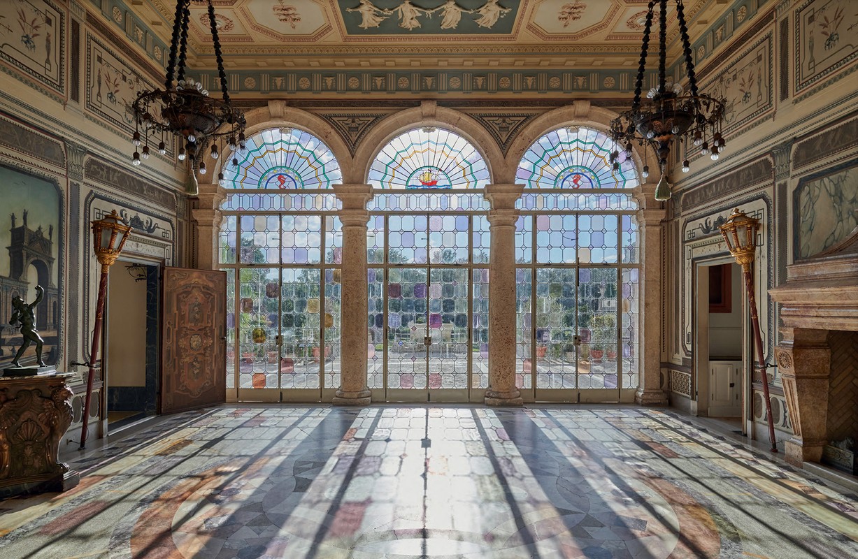 Grand interior view of a historic building featuring arched windows, marble floors, and ornate chandeliers, creating an elegant, sunlit space.