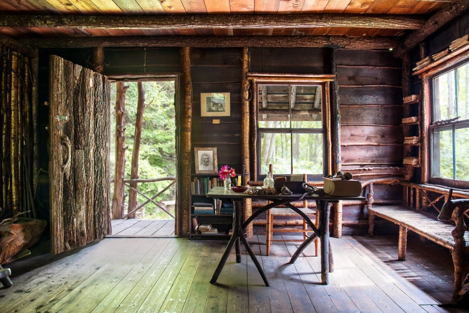 Rustic cabin interior with wooden walls, vintage furniture, open doorway, and windows looking out to a lush forest. Natural light fills the room.