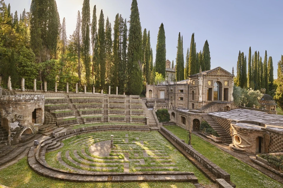 Outdoor view of an amphitheater with tiered seating, surrounded by tall trees and classical architecture in a serene park setting.