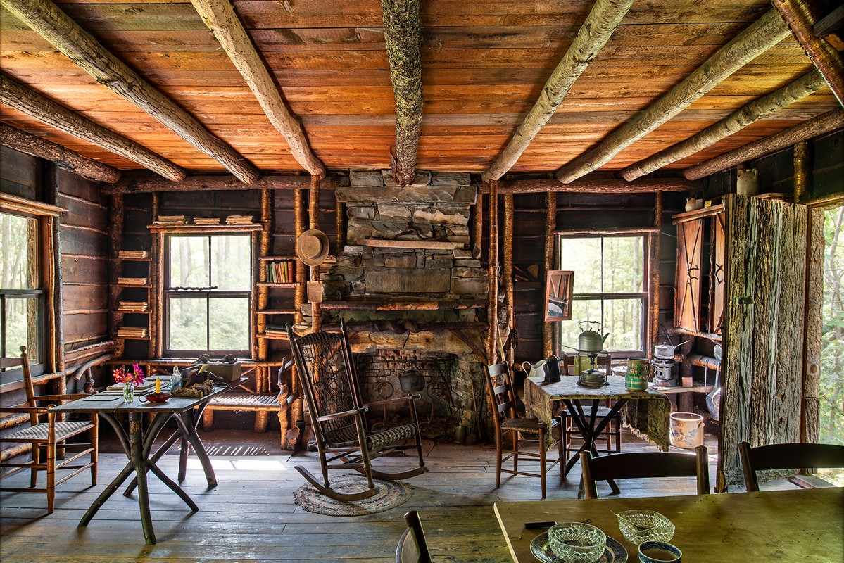 Rustic cabin interior with wood walls and ceiling, stone fireplace, rocking chair, tables, and chairs in a warm, inviting setting.
