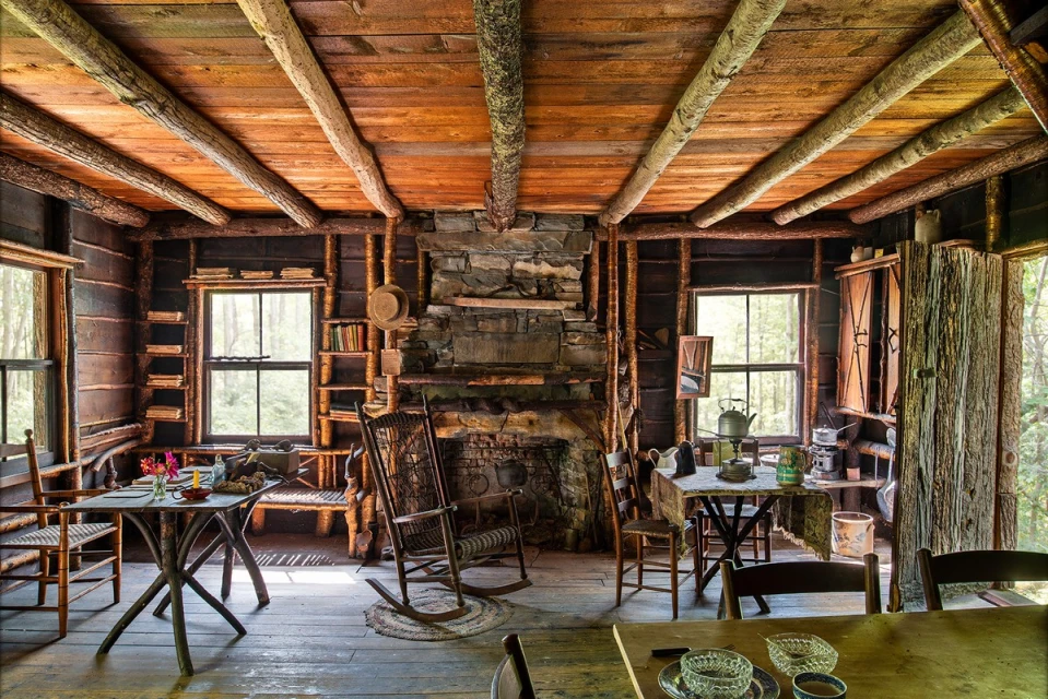 Rustic cabin interior with wood walls and ceiling, stone fireplace, rocking chair, tables, and chairs in a warm, inviting setting.