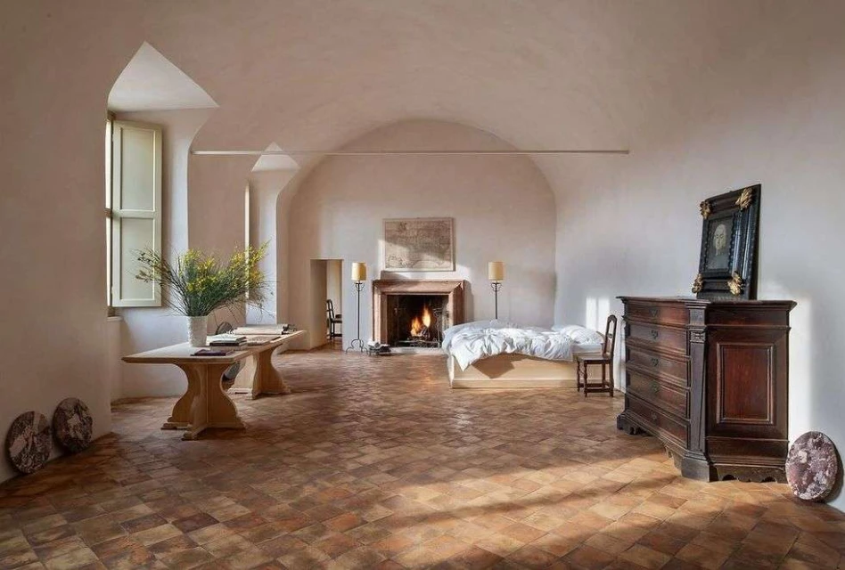 Interior bedroom boasts a fireplace, table with vase of yellow flowers, brick floors, and chest of drawers in a brightly lit space.