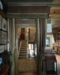 An ornate doorway draped with heavy brown curtains revealing a carpeted staircase inside an eclectic historic interior.