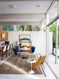 Modern interior living space with white walls, wooden cabinetry, blue chairs around a round table, and large glass windows.