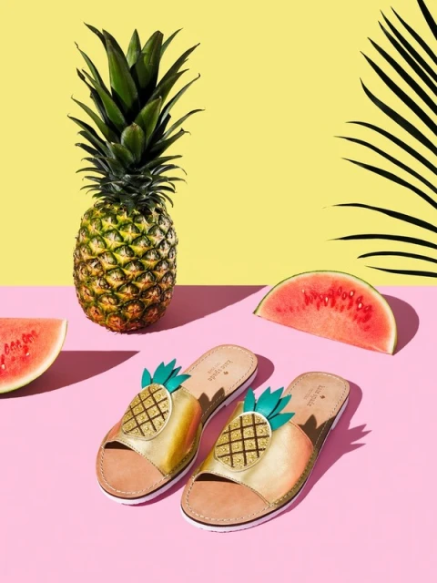Studio shot of Kate Spade pineapple sandals, a fresh pineapple, and slices of watermelon set against yellow and pink backgrounds.