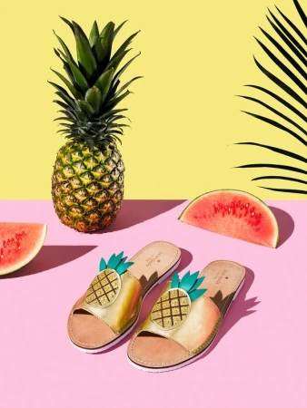 Studio shot of Kate Spade pineapple sandals, a fresh pineapple, and slices of watermelon set against yellow and pink backgrounds.