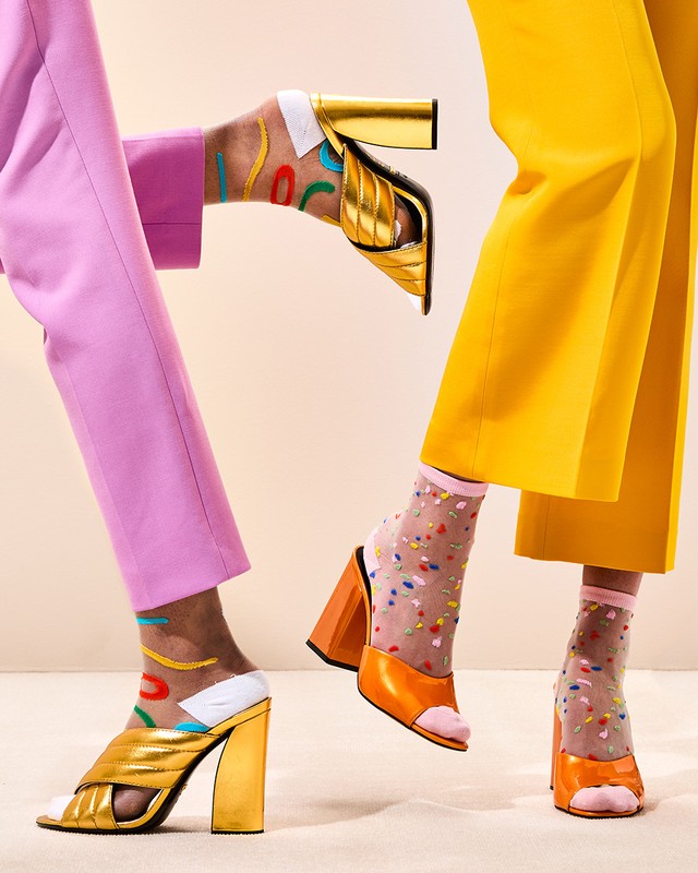 Fashion shot with two women's legs and feet wearing colorful socks, gold block-heeled shoes, yellow and pink pants on a light background.