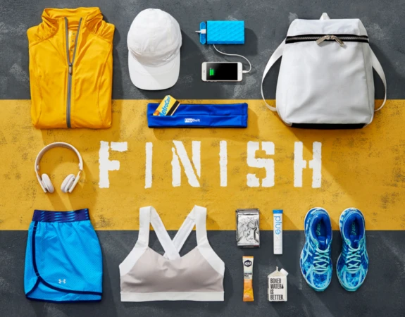 Overhead shot of athletic gear including athletic shoes, clothing, headphones, phone, portable charger, and snacks on yellow and gray background.
