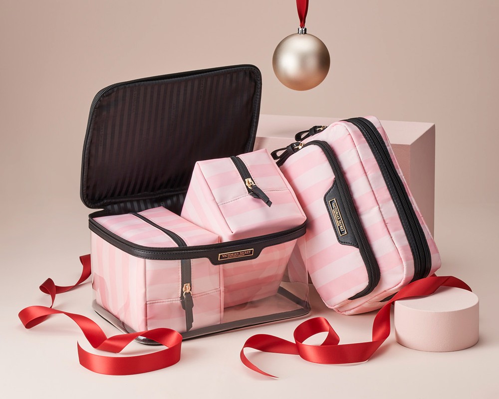 Still life of striped cosmetic bags and organizer case with red ribbon and gold Christmas ornament against a gradient background.
